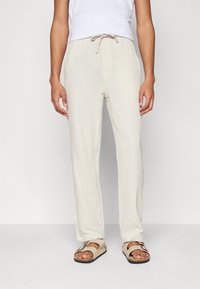 Persoon gekleed in lichtbeige, gestructureerde broek met trekkoord en beige open te sandaal met gespen, staand tegen een effen witte achtergrond.