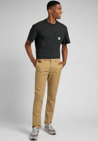 Mustat puuvillaiset t-paidat, joissa on tasku, yhdistettynä khakivihreisiin puuvillahousuihin. Henkilö seisoo rennosti, jalassaan harmaat ja valkoiset lenkkarit. Taustana on harmaa.