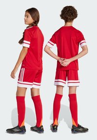 Tenue de football rouge avec des accents blancs, comprenant des manches courtes et un short. Comprend des chaussettes rouges arrivant aux genoux avec un dégradé orange et des chaussures à crampons noires.