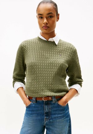 Olive green cable-knit sweater with a round neck, paired with a white collared shirt, denim jeans, and a brown belt.