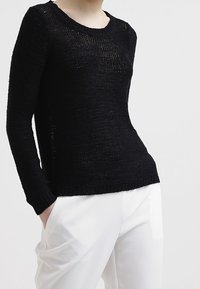 Schwarzer Strickpullover mit langen Ärmeln, strukturiertem Stoff, Rundhalsausschnitt und lockerer Passform, kombiniert mit weißen Hosen.