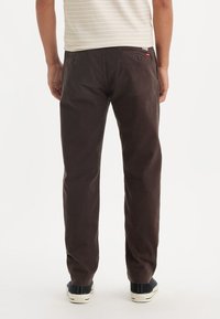Pantalon en coton marron foncé à coupe droite, avec des poches arrière boutonnées et une texture subtile. Porté avec des baskets bleu marine.