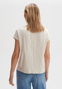 Nainen, jolla on olkapäivän pituinen vaaleanblonde hius, pukeutuneena vaaleaan teksturoituun lyhythihaiseen paita ja sinisiin farkkuihin, kääntyneenä pois valkoista taustaa vasten.