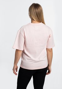 Lys rosa bomulls t-skjorte med korte ermer, med hevet tekst på baksiden. Klippet i en avslappet passform, den er kombinert med svarte bukser.