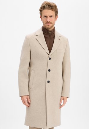 Mann mit hellbraunen Haaren und Bart, der einen beigefarbenen Drei-Knopf-Mantel und einen braunen, mit Reißverschluss versehenen Pullover mit hohem Kragen trägt, steht vor einem weißen Hintergrund.