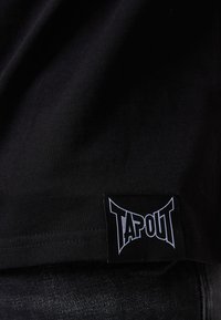 Camiseta de algodón negra con una textura suave, que presenta una prominente etiqueta del logo TAPOUT cosida en el dobladillo. La tela es suave y ligera.