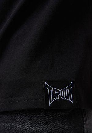 Sort bomulds T-shirt med en blød tekstur, prydet med en fremtrædende TAPOUT-logo label syet i kanten. Stoffet er glat og let.