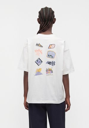 Personne aux cheveux tressés portant un T-shirt blanc oversize avec plusieurs logos colorés New Balance dans le dos, assorti à un pantalon foncé.