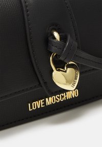 Borsa a tracolla nera in ecopelle con finitura testurizzata, dotata di un charm a forma di cuore dorato e accento con logo. Hardware argentato con un design elegante.