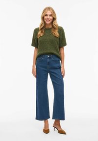 Jersey de color verde oliva de punto con mangas cortas, combinado con unos vaqueros azules de pierna ancha y cintura alta. Unos tacones marrones de punta afilada completan el conjunto.