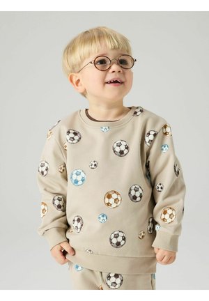 Ungt barn med blondt hår og briller, iført beige genser og bukser med trykk av flerfargede fotballer, smilende og ser oppover.