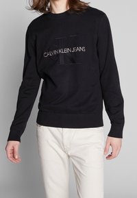 Pull en tricot noir avec poignets et ourlet côtelés, présentant un logo en relief "CK" et "CALVIN KLEIN JEANS" brodés en blanc contrastant.