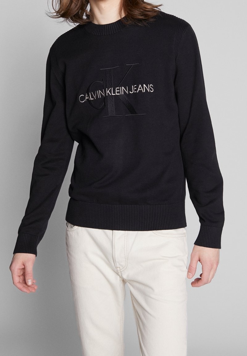 Pull en tricot noir avec poignets et ourlet côtelés, présentant un logo en relief "CK" et "CALVIN KLEIN JEANS" brodés en blanc contrastant.
