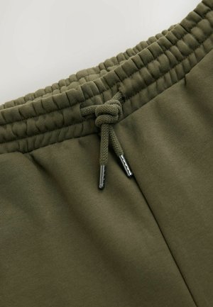 Pantaloni jogger verde oliva con fascia elastica in vita, dotati di un cordino intrecciato con punte in metallo. Tessuto morbido e vestibilità rilassata.
