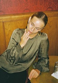 Olivgrön satinblus med puffärmar och rynkade manschetter. Modell som vilar hand på hakan, sittande vid ett bord med glas.