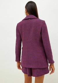 Blazer en tweed violet avec tissage texturé, col cranté et manches effilochées ; associé à une jupe assortie, mettant en valeur un design coordonné.