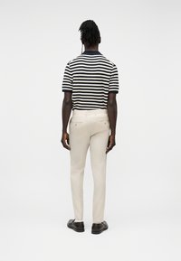 Homme debout, de dos, portant une chemise à manches courtes rayée noir et blanc, un pantalon beige clair et des chaussures marron foncé sur un fond uni.