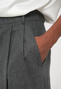 Pantaloni gri din lână cu pliuri, având un buzunar lateral. O mână este sprijinită în buzunar. Țesătura are un aspect moale și texturat.