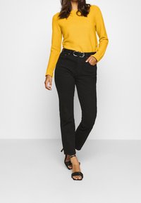 Top jaune à manches longues côtelé associé à un jean noir taille haute et des sandales noires à lanières. Comprend une ceinture avec une boucle argentée.