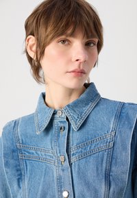 Lyseblå denimskjorte med spiss krage, sølvknapper og to horisontale sømmer over brystet. Glatt, teksturert stoff.