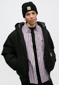 Črna puhovka s sijočim podlogom, oblečena čez svetlo vijolično in belo črtasto srajco. Črna kapa s logotipom spredaj.