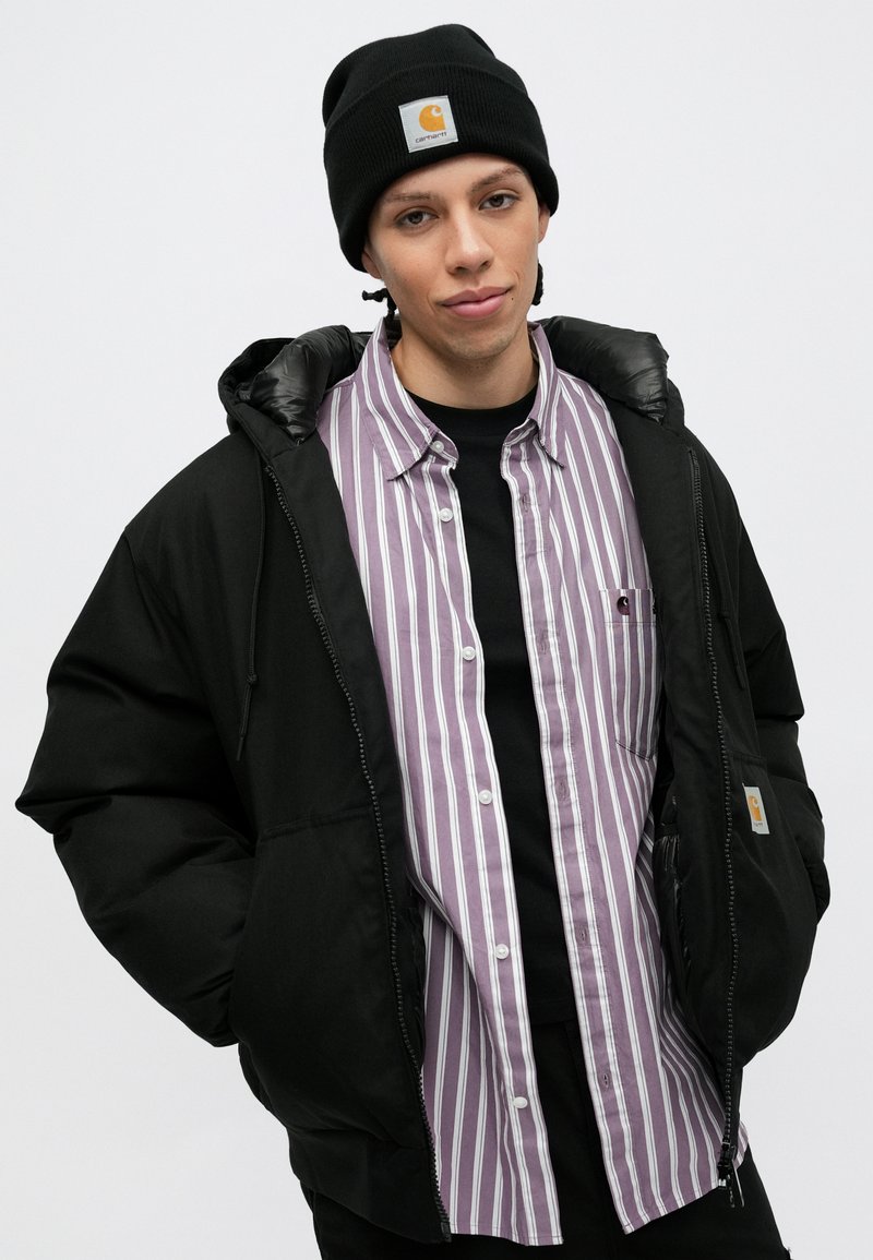 Chaqueta acolchada negra con un forro brillante, llevada sobre una camiseta a rayas de color púrpura claro y blanco. Gorro negro con un logo en la parte frontal.