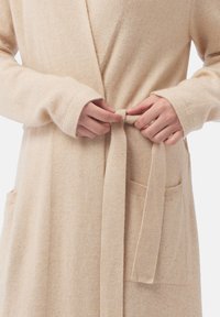 Un peignoir en tricot beige présente un design enveloppant avec une fermeture à nouer, des manches longues et deux poches avant. Texture douce et matériau léger.
