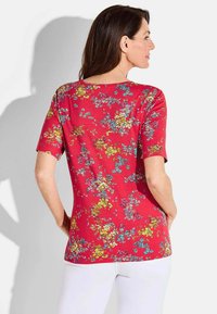 Rotes T-Shirt mit kurzen Ärmeln, das eine Vielzahl kleiner, bunter Blumen in Blau, Gelb und Pink auf einem lebhaften Hintergrund zeigt.