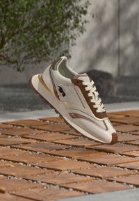 Beige og brun joggesko med nettingoverdel, kontrasterende detaljer og en teksturert gummisåle. Dekorativ logo på siden.