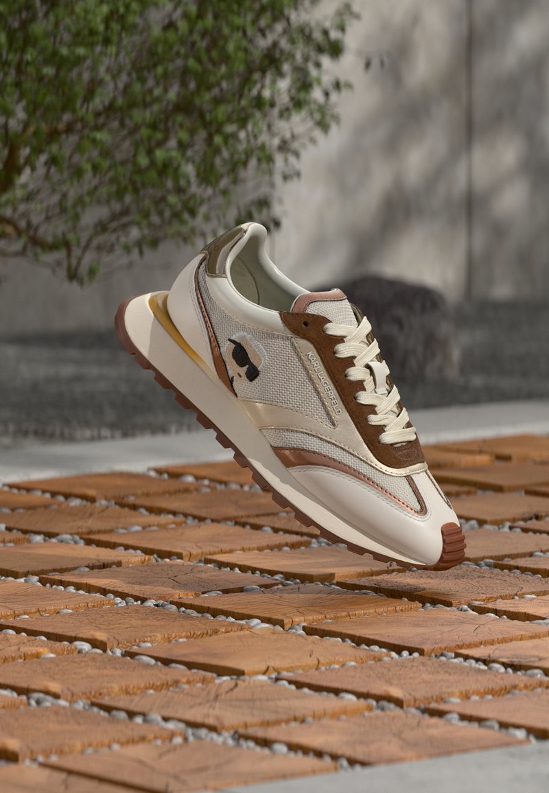 Beige og brun joggesko med nettingoverdel, kontrasterende detaljer og en teksturert gummisåle. Dekorativ logo på siden.