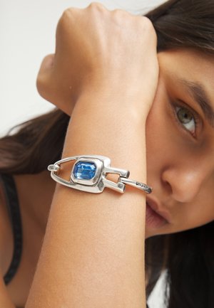 Femme reposant son bras levé sur son visage, portant un bracelet en argent avec une grosse pierre précieuse bleue à son poignet.