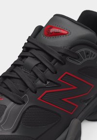 Κοντινό πλάνο αθλητικού παπουτσιού σε μαύρο και γκρι με κόκκινες λεπτομέρειες, μαύρα κορδόνια και ετικέτα "9060" στη μαξιλαρωμένη γλώσσα.