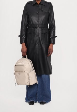 Zwarte lederen trenchcoat met knopen en zakken, met een ceintuur in de taille. Beige nylon rugzak met ritsen, gedragen met wijd uitlopende spijkerbroek.