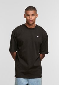 T-shirt nera oversize in cotone con maniche corte e collo a giro. Presenta un piccolo logo rosa sul petto. Design semplice.
