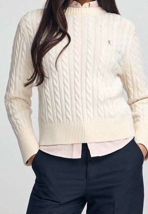 Vrouw draagt een crèmekleurige kabelgebreide trui over een roze geruit overhemd, gecombineerd met donkere broek, handen in de zakken, lang donker haar zichtbaar.