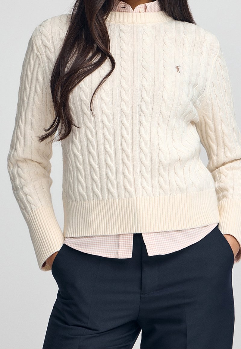 Femme portant un pull crème en maille torsadée par-dessus une chemise à carreaux rose, associée à un pantalon foncé, mains dans les poches, cheveux longs et foncés visibles.