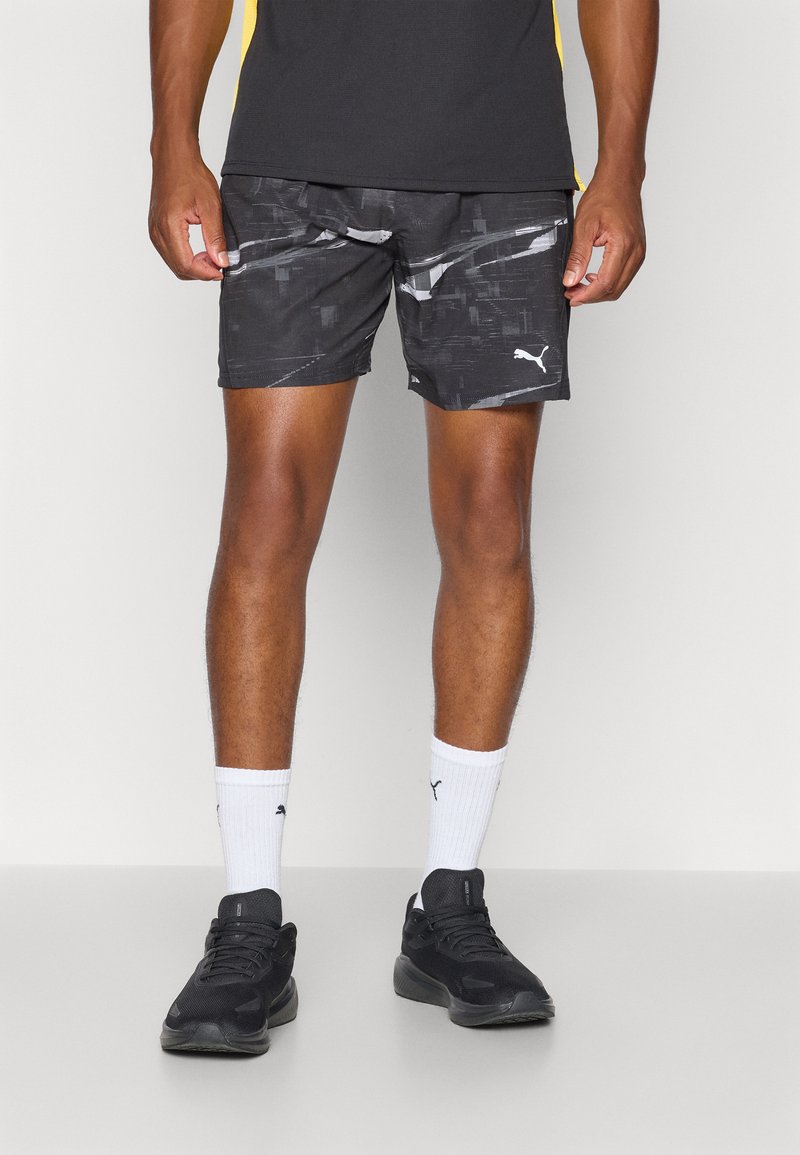 Schwarze Sportshorts mit einem geometrischen Muster, aus weichem Stoff, mit elastischem Bund und einem kleinen weißen Logo. Getragen mit weißen Socken und schwarzen Schuhen.