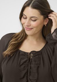 Mujer con cabello largo castaño, que lleva una blusa marrón oscuro con volantes, tocándose el cabello cerca de la oreja con un pequeño aro visible.