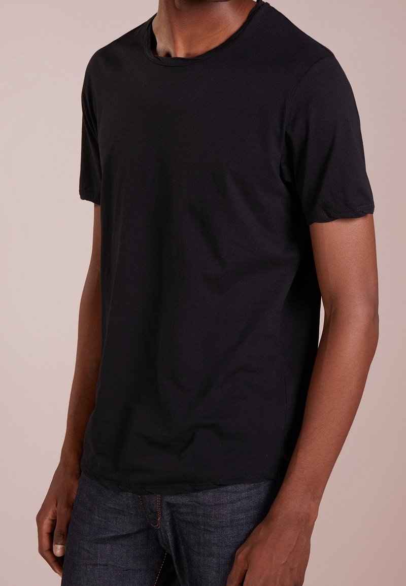 Homme portant un t-shirt noir uni à manches courtes et un jean bleu foncé, debout devant un fond beige clair.