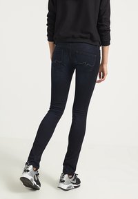 Mörka, stretchiga denimjeans med en slät yta, som har bakfickor med vågmönster och dekorativa nitar. Kombineras med sportskor.