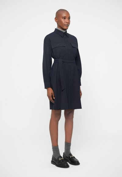 Μπλε πουκάμισο φόρεμα σε μεσαίου μήκους σχέδιο, με ώμους τσέπες, ζώνη που δένει και κουμπιά. Μαύρες loafers και γκρι κάλτσες ολοκληρώνουν την εμφάνιση.