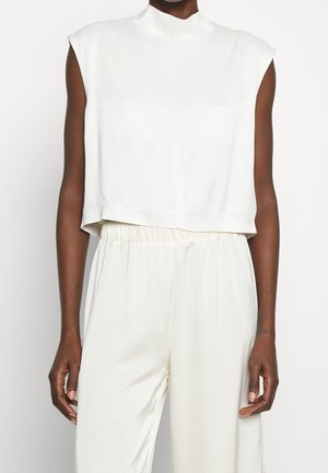 Haut court blanc sans manches à col montant associé à un pantalon blanc ample à taille élastique sur une personne à la peau foncée, sur fond uni.
