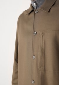 Camisa marrón de botonadura con cuello puntiagudo, que cuenta con un bolsillo en el pecho y una capa gris con cremallera debajo, textura de tela suave.