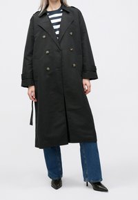 Gabardina negra de doble botonadura con botones dorados, mangas largas y cintura con cinturón, combinada con vaqueros azules de pierna ancha y tacones negros puntiagudos.