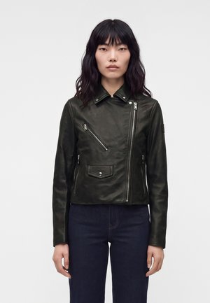 Femme aux cheveux noirs mi-longs portant une veste en cuir noire ajustée avec une fermeture éclair diagonale et un jean bleu foncé, debout devant un fond uni.