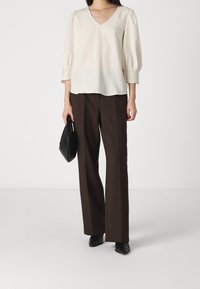 Blusa ecrù con maniche a sbuffo e bordo in pizzo, abbinata a pantaloni marroni a vita alta e una borsa nera, su uno sfondo semplice.