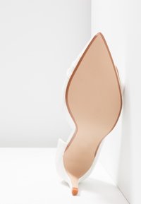 Chaussure blanche à talons hauts avec bout pointu, posée en angle contre un coin blanc, montrant la semelle beige lisse et le petit embout du talon.