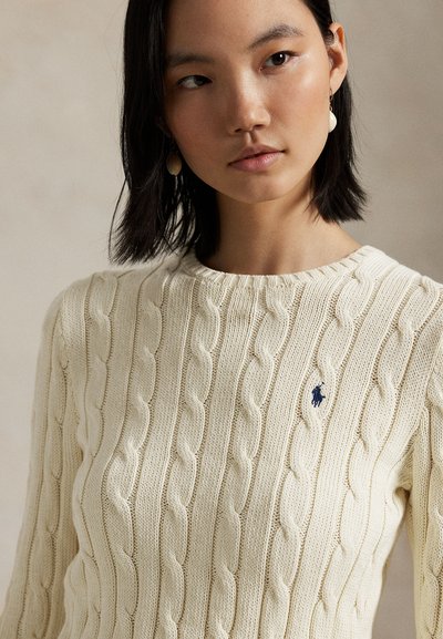 Jeune femme aux cheveux noirs porte un pull en maille torsadée crème avec un logo bleu brodé et des boucles d'oreilles pendantes en perles sur fond neutre.
