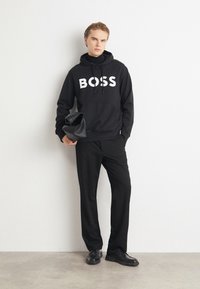 Juodas džemperis su "BOSS" logotipu, laisvas kirpimas, su raišteliais, derinamas su pritaikytais juodais kelnėmis ir blizgiais juodais batais. Laiko juodą rankinę.