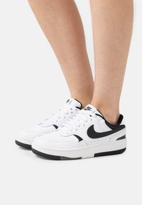White leather sneakers with black accents, round toe, perforated detailing, laces, and a textured black rubber sole. Nike branding on tongue and side.